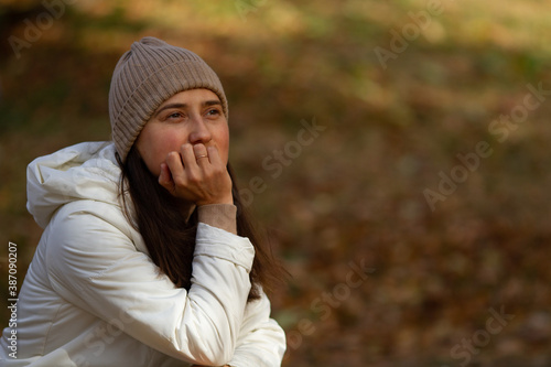 Pretty brunette woman sitting in the park thinking on a beautiful sunny autumn day. Woman wearing knitted hat, warm jacket. Meditative mood. Horizontal shot. Copy space 