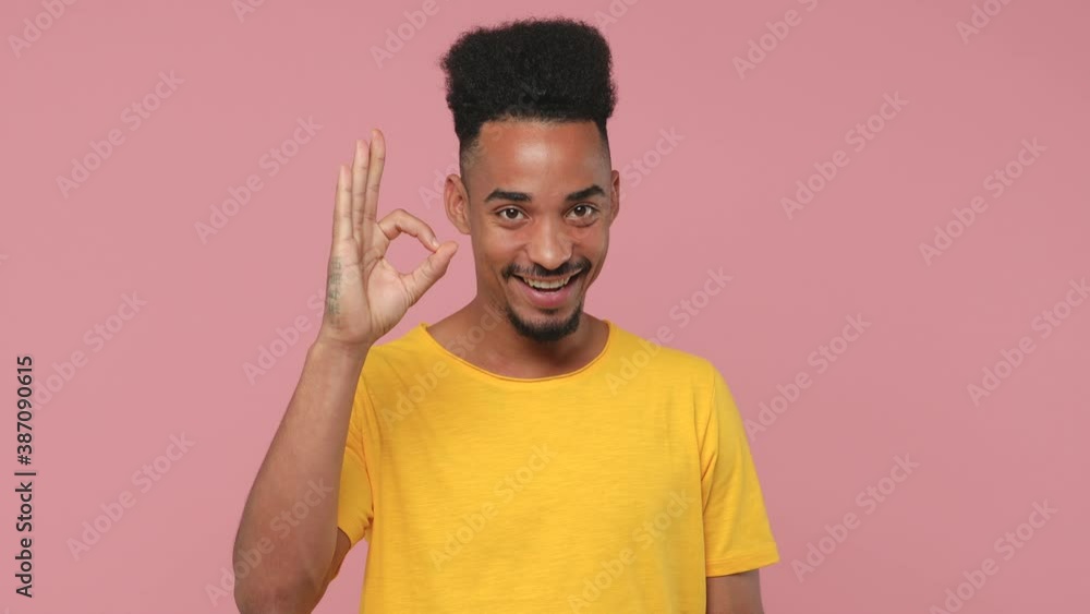 Excited cheerful young african american man 20s years old in yellow t-shirt posing isolated on pastel pink background in studio. People lifestyle concept. Looking camera say wow showing OK gesture Excited cheerful young african american man 20s years old in yellow t-shirt posing isolated on pastel pink background in studio. People lifestyle concept. Looking camera say wow showing OK gesture