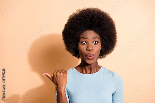 Photo portrait of impressed african american woman pointing thumb at blank space isolated on pastel beige colored background