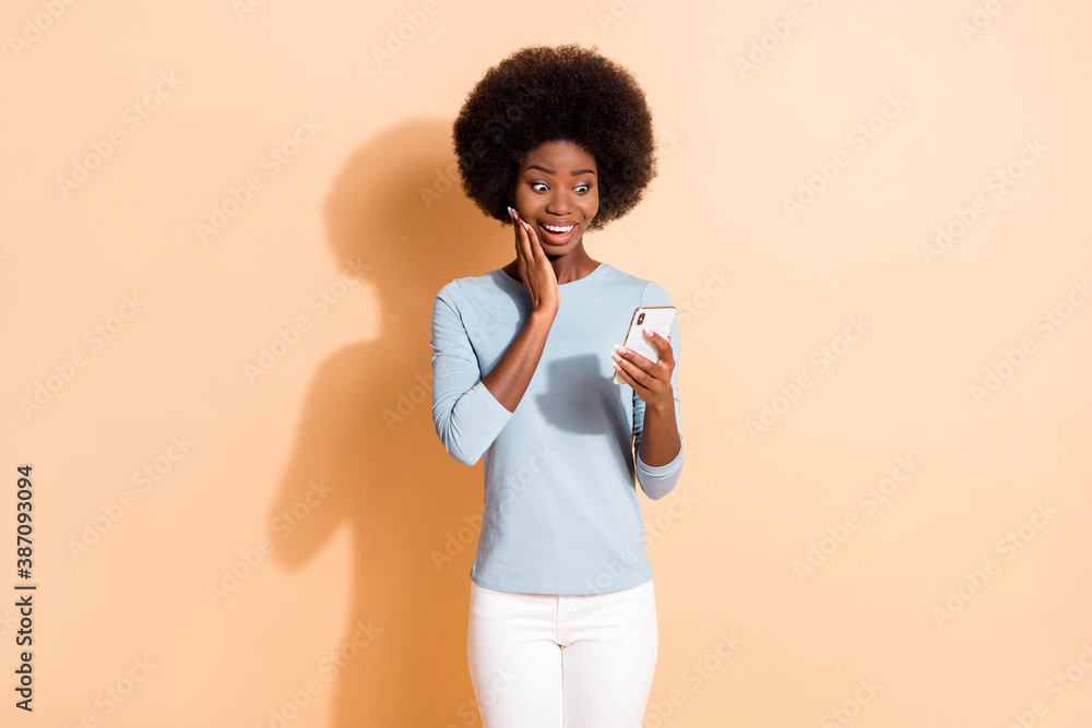 Photo portrait of shocked impressed dark skinned girl reading information from smartphone touching face with hand isolated on beige color background - obrazy, fototapety, plakaty 