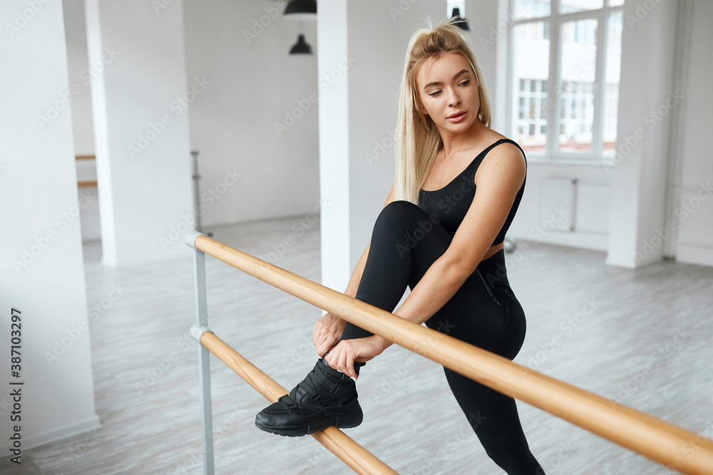 Beautiful sports model woman with a slender body in fashionable sportswear does stretching of the legs in a bright room. blonde in black suit warming up Beautiful sports model woman with a slender body in fashionable sportswear does stretching of the legs in a bright room. blonde in black suit warming up
