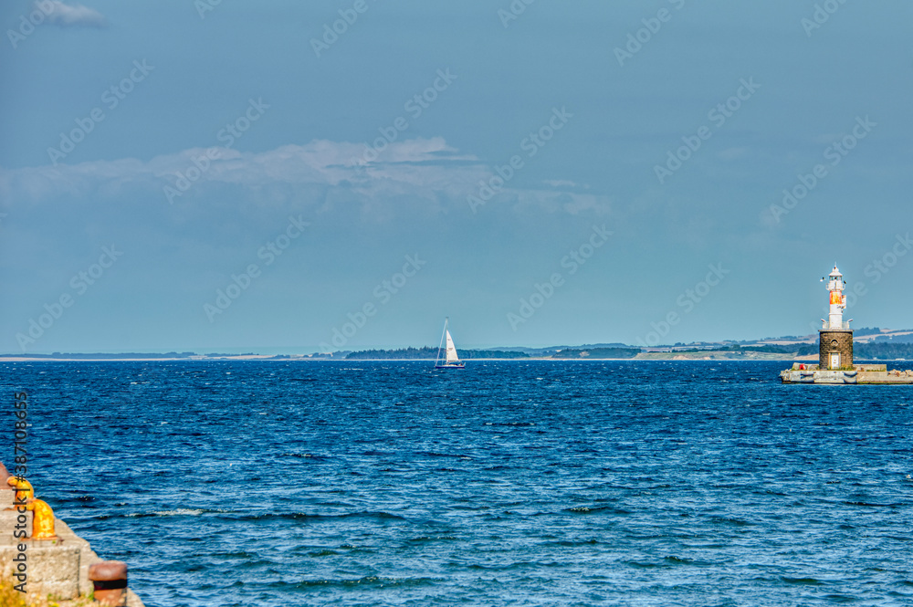 A lonely sailing boat in Aarhus harbour sails in a calm Baltic sea beside Arhus Lighthouse inspiring a romantic escape or sport activity. Illustration for sailing module or sail training - Denmark