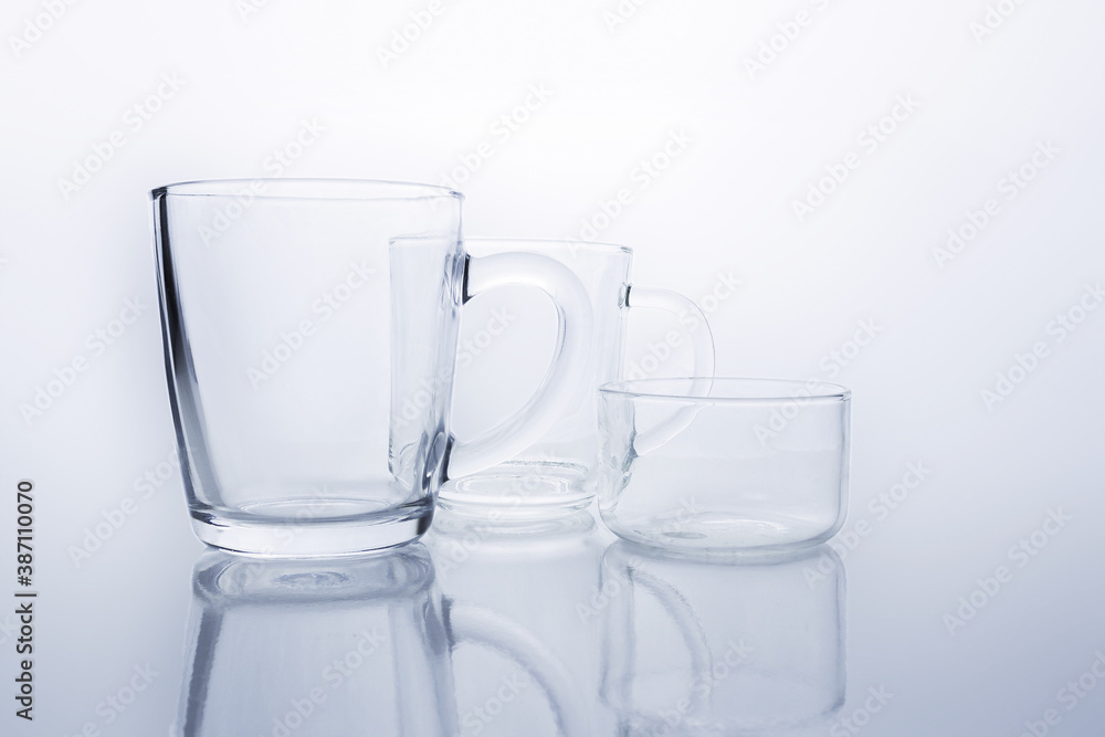 Two empty transparent glass mugs and saucer rosette on reflective surface on white background. Transparent glass still life. Two empty transparent glass mugs and saucer rosette on reflective surface on white background. Transparent glass still life.