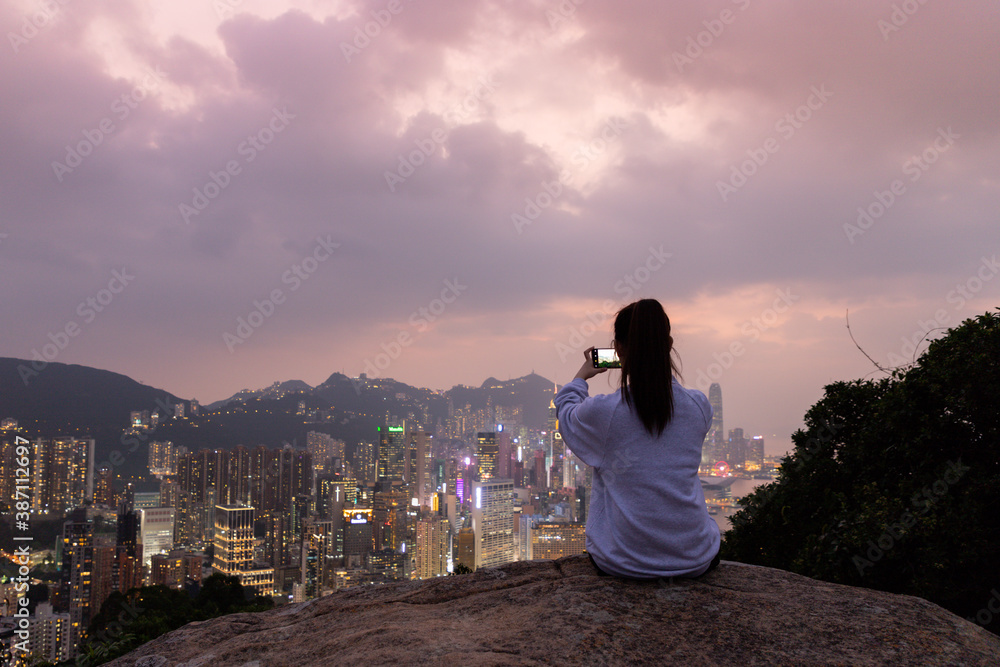 Fototapeta premium Silhouette of a girl who sits at rock and takes photos of sunset night view of city Skyline of Hong Kong Island (south of Victoria Harbour) from Braemar hill, North Point