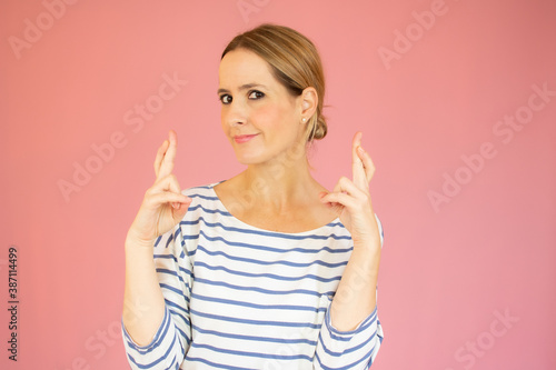 Tableau sur toile Indoor shot of glad happy woman wears striped t-shirt, smiles broadly, keeps fingers crossed, hopes for good luck, isolated against pink background with copy space for your text