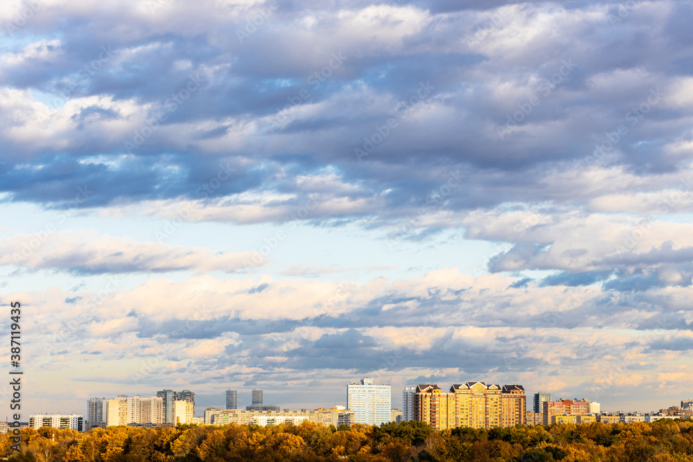 blue sky with low dark blue clouds over urban park and residential district on horizon lit by autumn sunset sun in Moscow city
