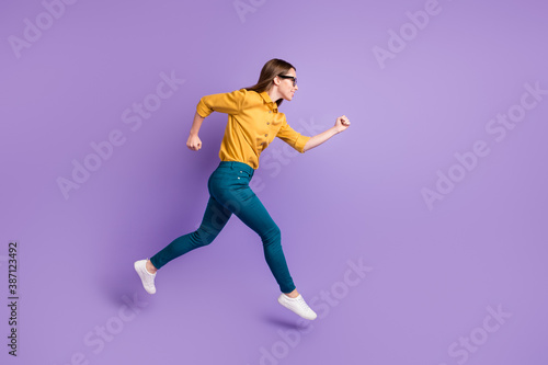 Full length profile photo of girl run jump hurry wear yellow shirt blue trousers sneakers isolated purple color background