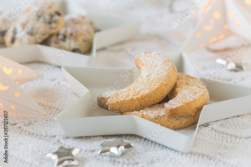 Vanilla-flavored crescent cookies on a white star plate on white blanket, decorated with silver stars and starlight, mini stollen in the background