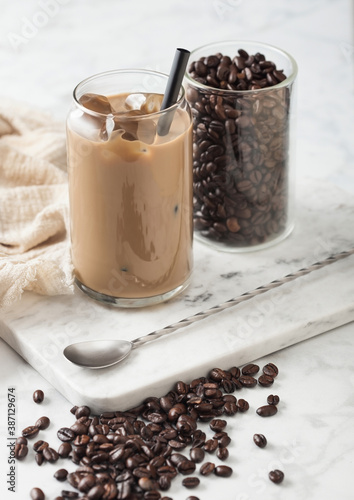 Iced cold caramel coffee with milk and glass container of beans and spoon on marbel board and light background.