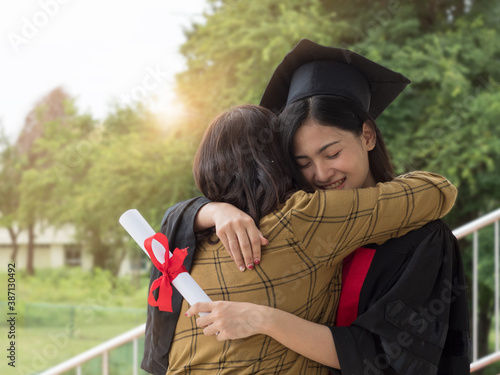 Asian female graduate wearing mortar board, holding certificate hugging senior woman tight with happiness.