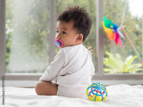 Cute little Asian baby boy sitting on the white soft blanket with pacifier in the mouth.