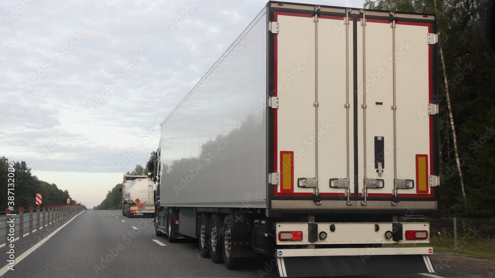 European road semi trucks with white trailer van and barrel move on two lane suburban asphalted highway motorway, rear view at summer evening on forest and sky background, delivery cargo logistics