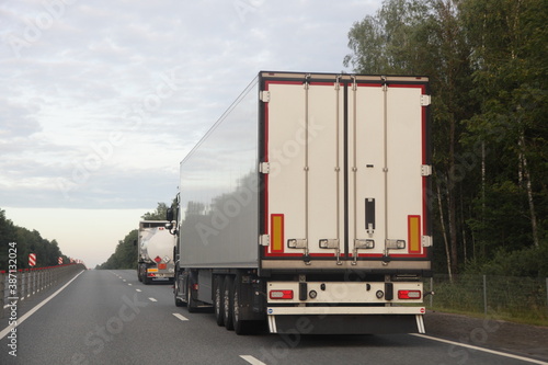 European van truck with white semi trailer and fuel truck drive on two lane suburban asphalted highway road, back view at summer evening on forest and sky background, transportation logistics