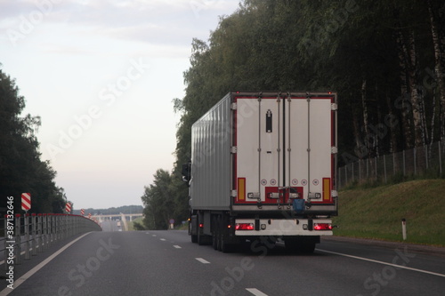 European long vehicle truck with white semi trailer van move on two lane suburban asphalted highway road, back view at summer evening on forest and sky background, carriage transportation logistics