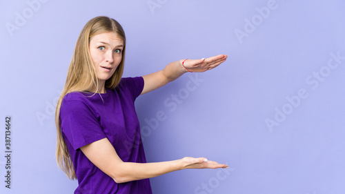 Young blonde woman isolated on purple background shocked and amazed holding a copy space between hands.