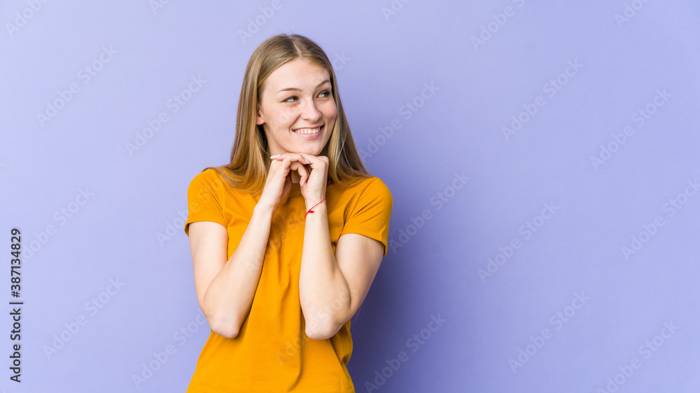 Young blonde woman isolated on purple background keeps hands under chin, is looking happily aside. Young blonde woman isolated on purple background keeps hands under chin, is looking happily aside.