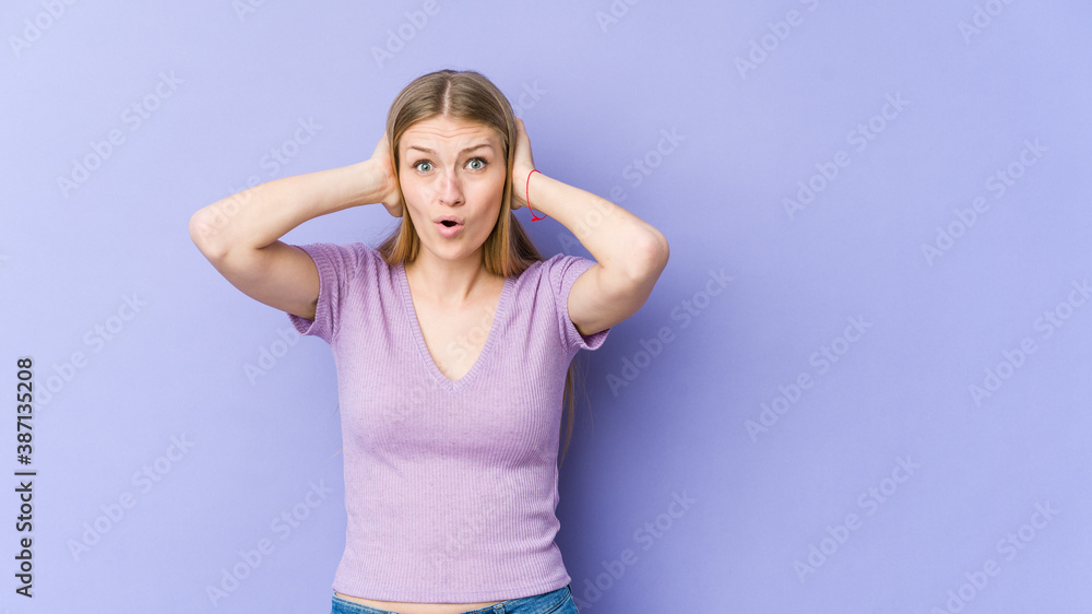 Young blonde woman isolated on purple background covering ears with hands trying not to hear too loud sound.