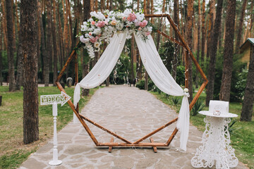 A wooden wedding arch decorated with flowers and white cloth stands in the forest among the pine trees in nature. Festive ceremony.