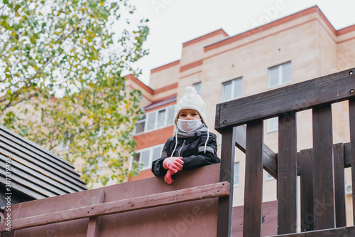 a 3-year-old child in a black overalls, a hat and a medical mask walks alone on the playground during the coronavirus pandemic in Russia 