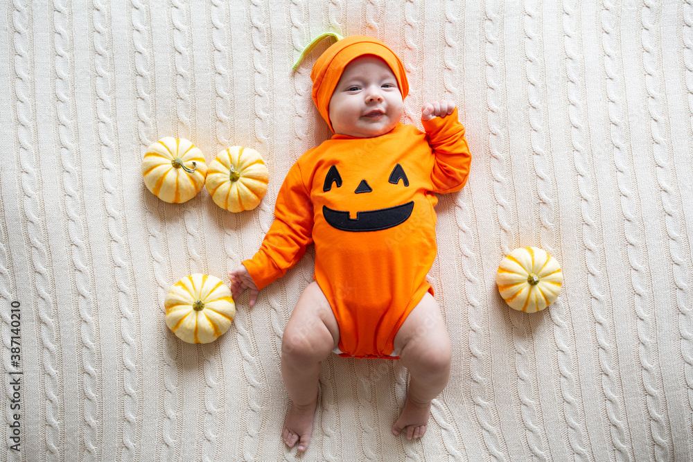 Fototapeta premium High Angle View Of Baby In Pumpkin Costume. Happy Kid two months old in orange Halloween costume on bed