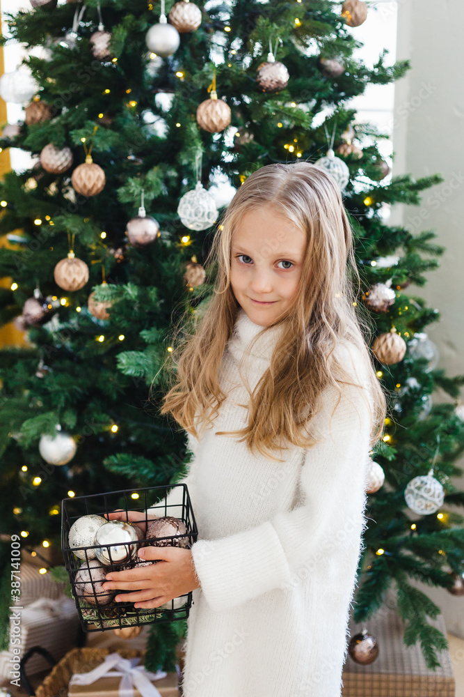 A girl stands near the Christmas tree and holds a basket with balloons for decorating the Christmas tree, long blonde hair, white sweater, baby, daughter A girl stands near the Christmas tree and holds a basket with balloons for decorating the Christmas tree, long blonde hair, white sweater, baby, daughter