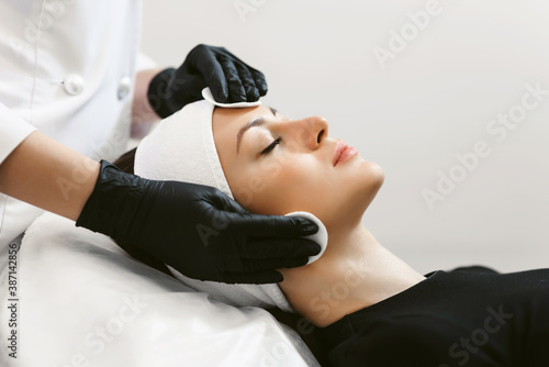 Close up of hands of skillful beautician cleaning and touching female face with cotton pad or sponge. The woman is lying and relaxing. Her eyes are closed with pleasure