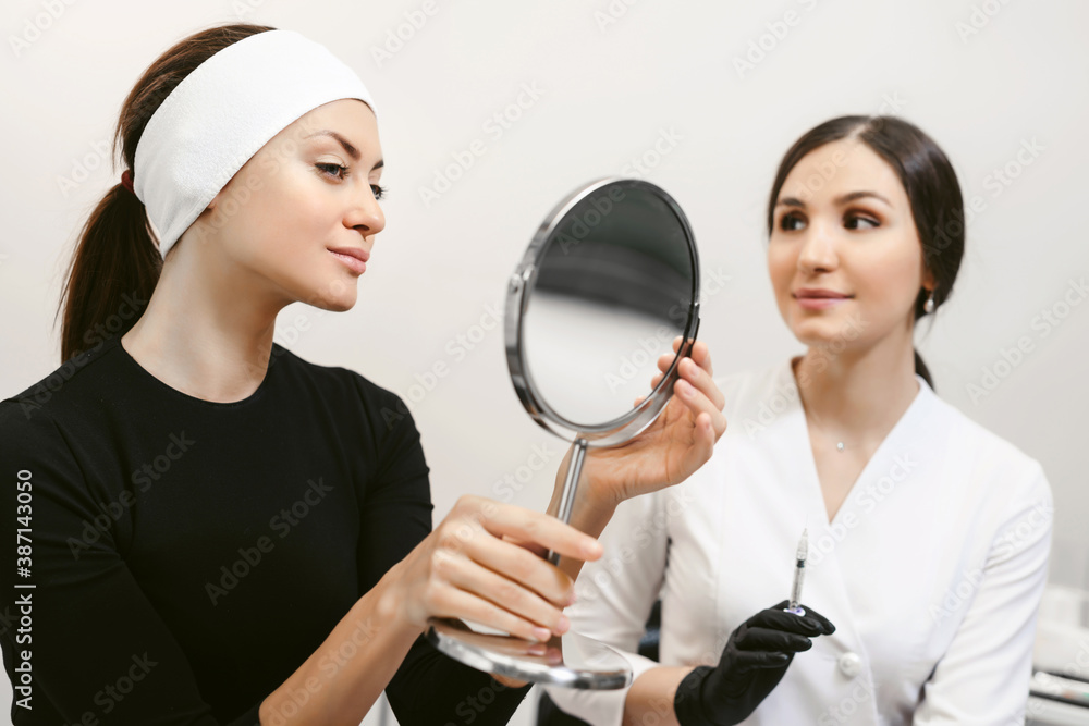 Beautiful young woman looking at herself in the mirror after cosmetic procedures in spa salon or beauty clinic. Smiling beautician doctor with syringe at background. Successfull cosmetology concept. Beautiful young woman looking at herself in the mirror after cosmetic procedures in spa salon or beauty clinic. Smiling beautician doctor with syringe at background. Successfull cosmetology concept.