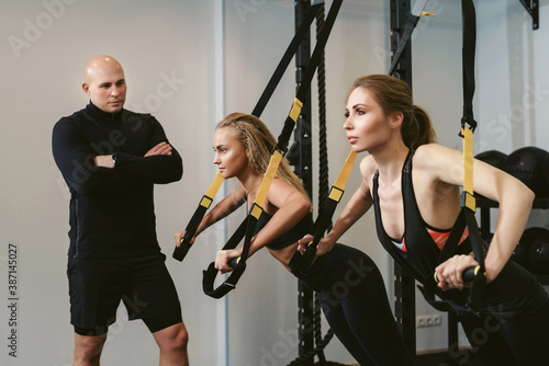 Two young women doing a suspension push ups training with their coach at a fitness club. Private workout in gym with a personal trainer. The concept of losing weight and gaining muscle mass.