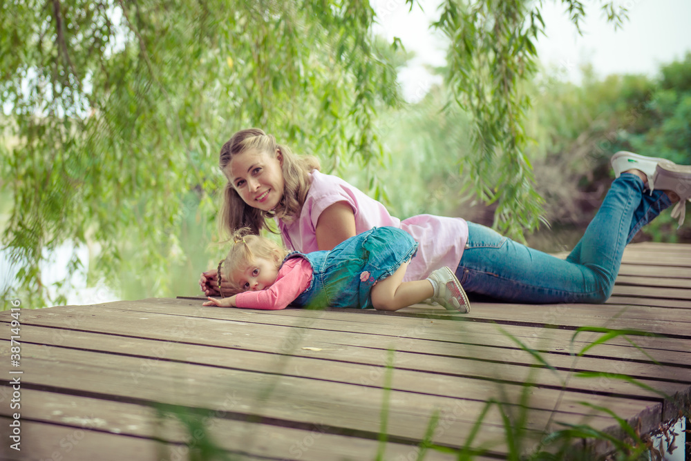 Fototapeta premium Baby with mom. Family values. Portrait of mom and daughter is sitting on a wooden bridge near the river. Mother and baby.