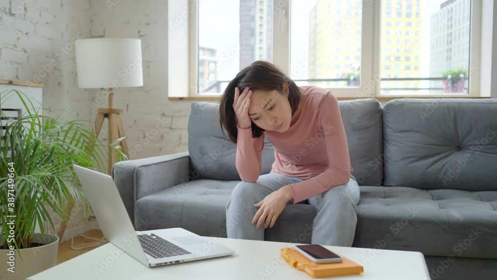 Depressed asian ethnicity young woman sitting on sofa, thinking of personal problems alone at home, unhappy tired mixed race lady suffering from mental stress and feeling sad Depressed asian ethnicity young woman sitting on sofa, thinking of personal problems alone at home, unhappy tired mixed race lady suffering from mental stress and feeling sad