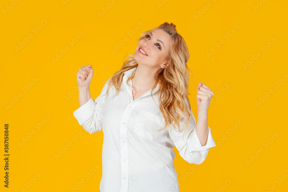 Excited happy European blonde woman wearing white shirt, enthusiastically shouts yes, chants and triumphs, becomes champion, wins the competition. Standing against yellow background. Excited happy European blonde woman wearing white shirt, enthusiastically shouts yes, chants and triumphs, becomes champion, wins the competition. Standing against yellow background.