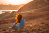 A young beautiful woman in jeans is sitting in the grass on a hillside in the orange rays of the sunset. Picturesque mountain landscape with the sea at sunset
