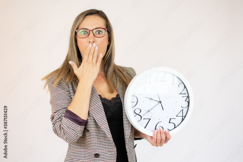 Young beautiful woman wearing jacket holding big clock covering mouth with hand, shocked and afraid for mistake. surprised expression Young beautiful woman wearing jacket holding big clock covering mouth with hand, shocked and afraid for mistake. surprised expression