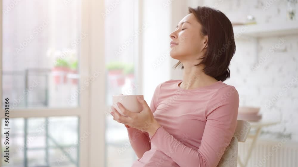 Mixed race satisfied asian female enjoying cup of coffee or tea drinking warm beverage at home. Young girl relaxing with morning drink in hands looking through window enjoying and dreaming Mixed race satisfied asian female enjoying cup of coffee or tea drinking warm beverage at home. Young girl relaxing with morning drink in hands looking through window enjoying and dreaming