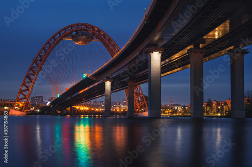View of Zhivopisny Bridge in Moscow, Russian Federation. Photo shoot of the red arch, steel cable-stayed bridge over the Moskva River. Evening. City lights and bridge lights are reflected in the water
