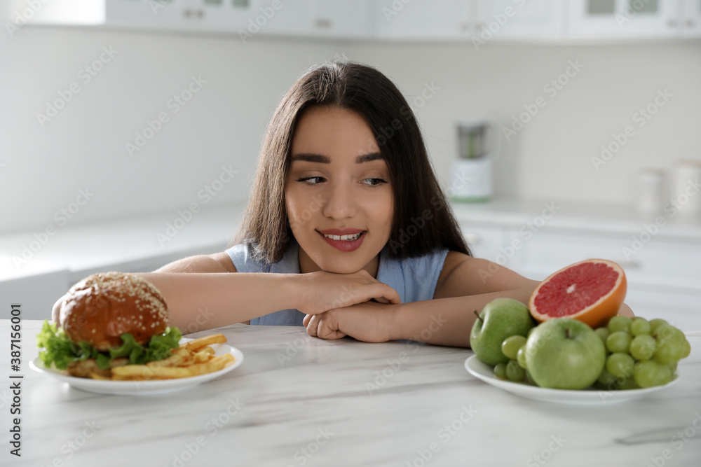 Woman choosing between fruits and burger with French fries in kitchen Woman choosing between fruits and burger with French fries in kitchen