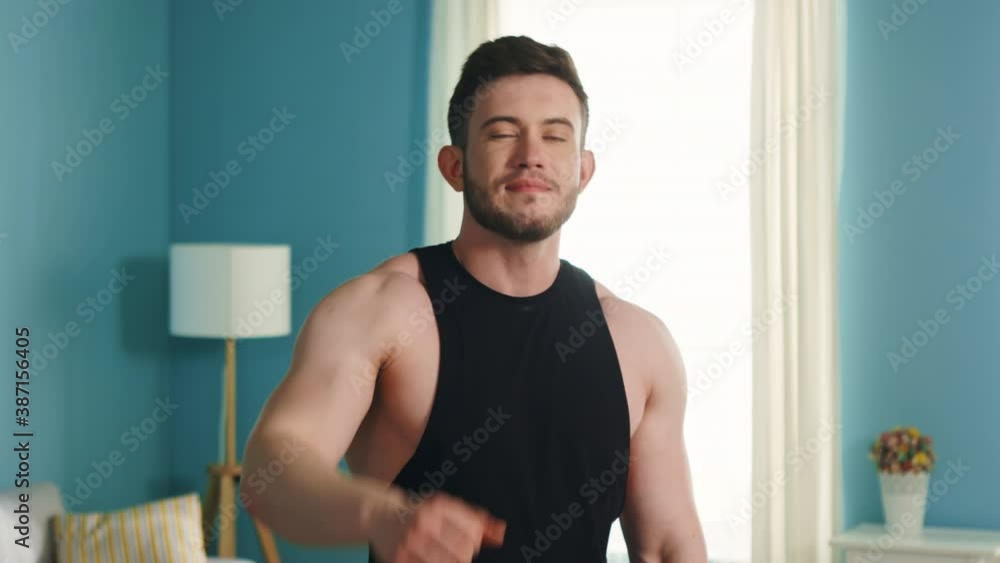 Portrait of young athletic man that is standing at home after daily workout, in sport outfit, happy and confident, smiles at camera and shows Portrait of young athletic man that is standing at home after daily workout, in sport outfit, happy and confident, smiles at camera and shows