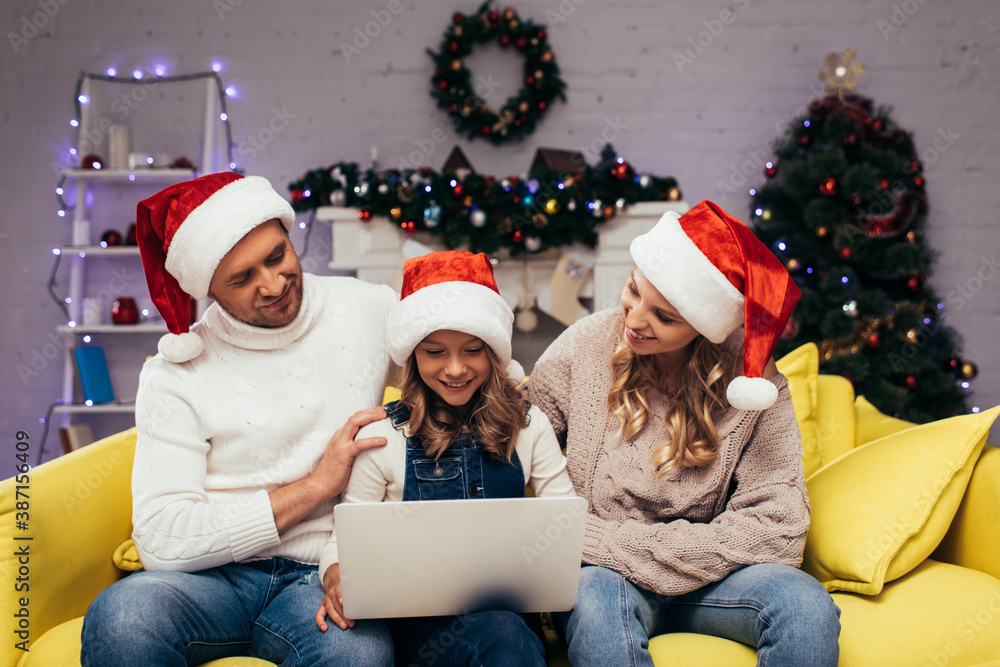 joyful kid in santa hat using laptop near parents in decorated living room joyful kid in santa hat using laptop near parents in decorated living room