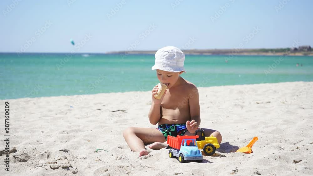 boy dressed in a Panama hat eats ice cream sitting on a sandy beach, against the background of a beautiful azure sea and blue sky on a Sunny day. boy dressed in a Panama hat eats ice cream sitting on a sandy beach, against the background of a beautiful azure sea and blue sky on a Sunny day.