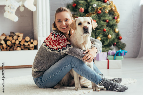 happy woman in sweater sitting on floor with labrador near decorated christmas tree