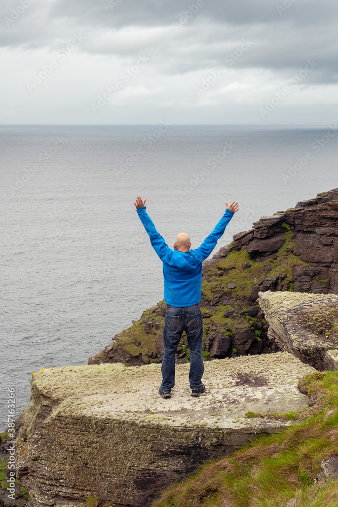 Bold male tourist standing on edge of Cliff of Moher, Ireland, Hands up in the air, back to the viewer.