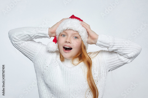 attractive red-haired teenage girl wearing Santa hat. she holds her head in her hands in amazement. photo session in the studio on a white background
