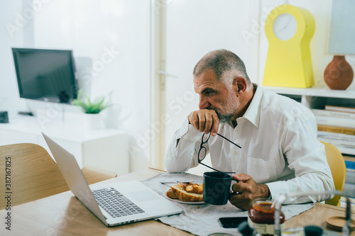 senior man having breakfast at home, using laptop computer