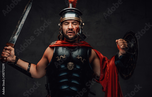 Brutal roman champion in dark armour with helmet and red cloak poses holding his sword and shield in dark background.