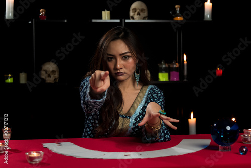 Portrait of Asian beautiful Gypsy fortune teller woman in dark room with white tarot card and pointing finger to camera. Focused on face.