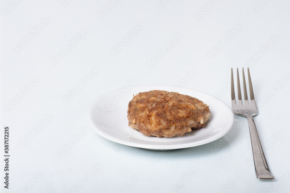 Cooked meat patty on a white plate next to a fork isolated on white background, close-up, copy space Cooked meat patty on a white plate next to a fork isolated on white background, close-up, copy space
