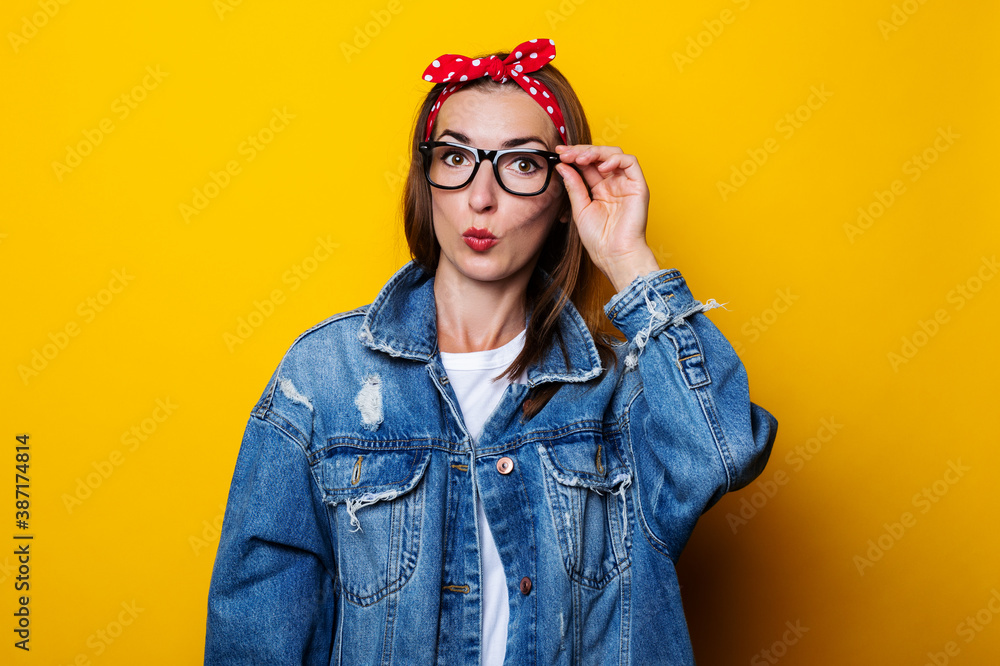 Young woman in hair band, in a denim jacket holding glasses making lips with a bow on a yellow background. Young woman in hair band, in a denim jacket holding glasses making lips with a bow on a yellow background.