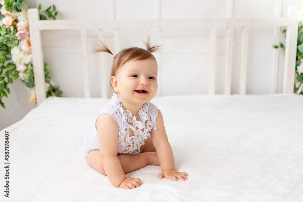 funny baby girl six months old sitting in a bright beautiful room on a white bed in a lace bodysuit looks away and smiles funny baby girl six months old sitting in a bright beautiful room on a white bed in a lace bodysuit looks away and smiles