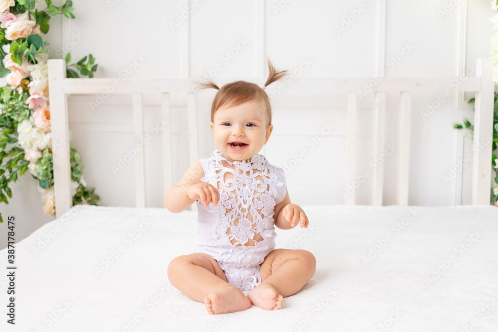 cheerful baby girl six months old sitting in a bright beautiful room on a white bed in a lace bodysuit and smiling cheerful baby girl six months old sitting in a bright beautiful room on a white bed in a lace bodysuit and smiling