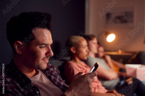 Man Using Mobile Phone Whilst Friends Watch TV At Home In Evening